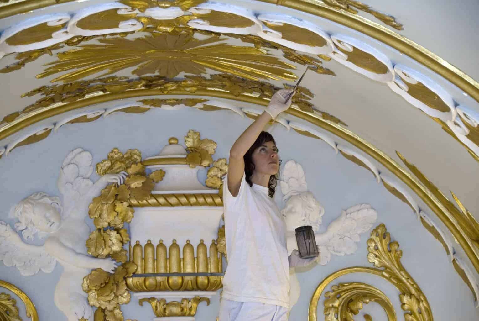 Restauradora de El Barco aplicando pan de oro en molduras ornamentales, proceso de restauración decorativa en techos con relieves y motivos clásicos